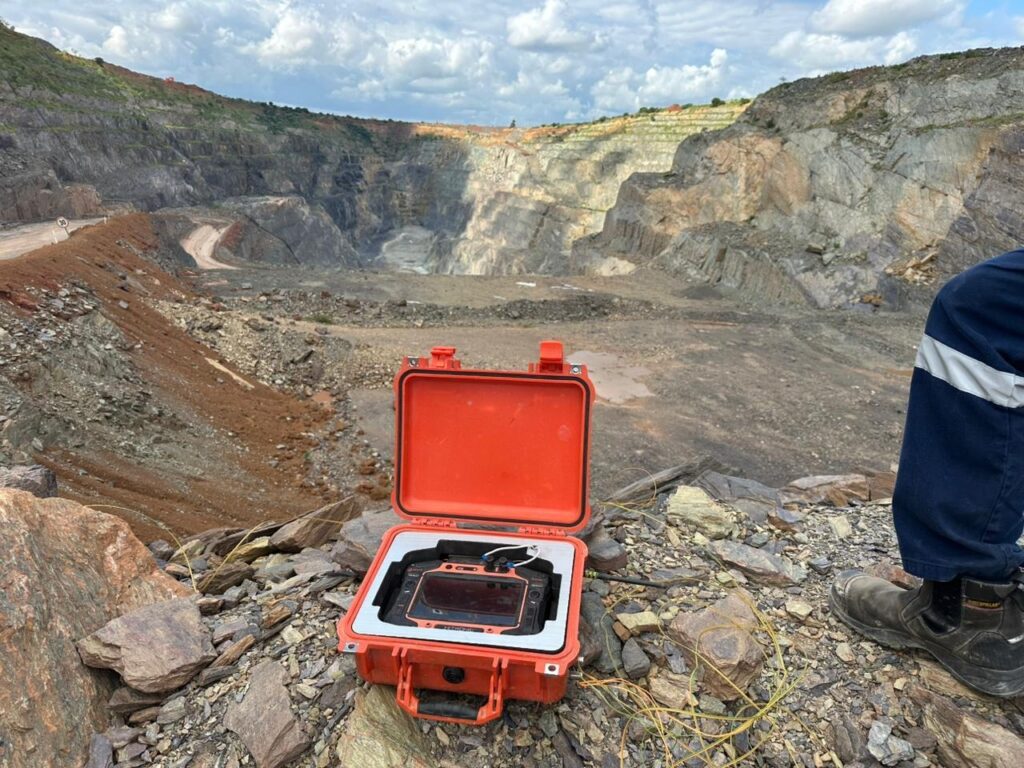 Blast monitoring equipment in an orange protective case on the pit rim, with an open-pit bench and haul road in the background.